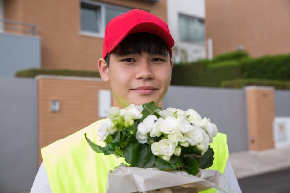A young male delivery person with light skin, wearing a red cap and a bright yellow safety vest, holds a bouquet of white flowers with green foliage wrapped in white paper. He stands outdoors in front