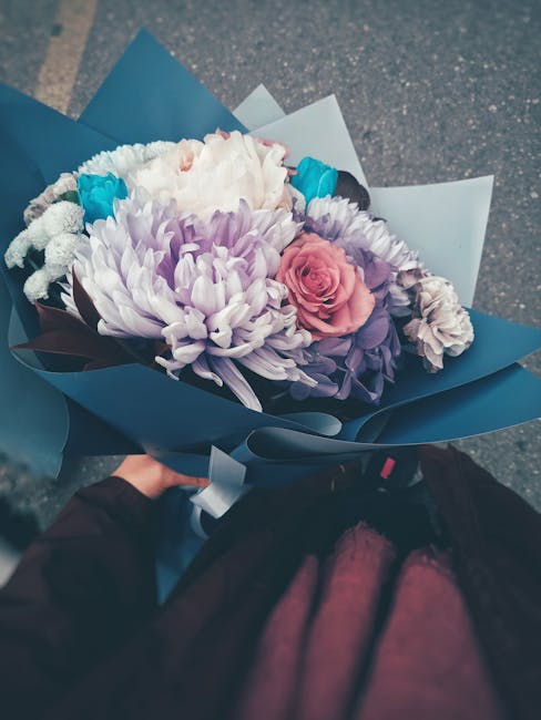 A hand holding a beautifully arranged bouquet featuring a large, white chrysanthemum at the center, surrounded by delicate pink roses, purple lisianthus, and small blue accent flowers. The bouquet is 
