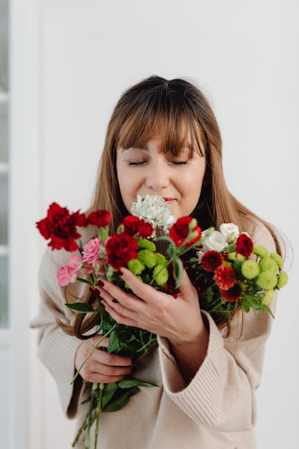 A woman with light brown hair and bangs, dressed in a beige knit sweater, is gently holding a vibrant bouquet of fresh flowers. The bouquet features a mix of red carnations, small pink roses, white hy