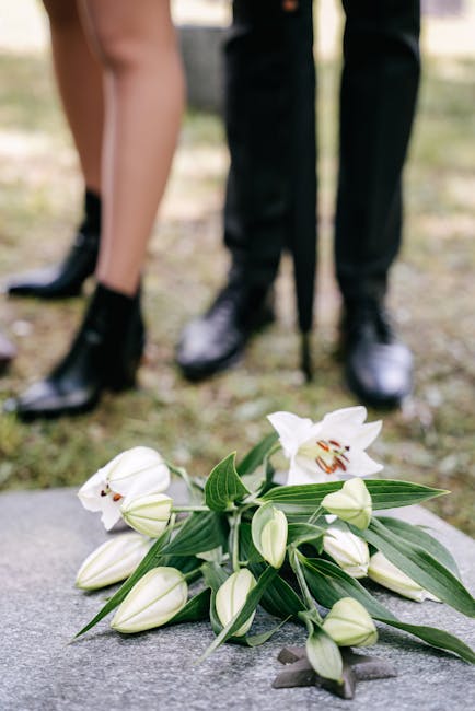 A bouquet of fresh white lilies with open blooms and closed buds, accompanied by long green leaves, lies on a stone surface. The flowers have smooth, curved petals with prominent orange stamens visibl