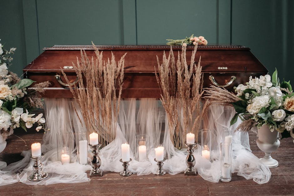 A wooden casket is decorated with elegant floral arrangements and candles for a funeral setting. The arrangement features soft white and blush roses, white orchids, and lush green foliage, placed in w