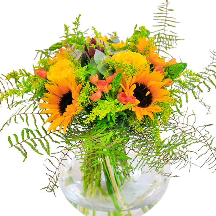From a local Cricklewood florist's hands comes this radiant bouquet, a vivid celebration of late summer's bounty reflected in every detail. The arrangement is abundantly set in a modern, spherical glass vase that reveals the sharply contrasting green stems and sparkling water within, grounding the design on a clean backdrop. Sunflowers, with their broad, amber petals and dark brown hearts, immediately draw the eye, bringing to mind the golden glow that spills over Cricklewood's parks in the evenings. Surrounding these, plush yellow roses unfurl in elegant, tightly layered coils, their velvety texture and sweet perfume lending a touch of classic English romance. Clusters of glossy red Hypericum berries add a lively pop of colour, standing out amid a generous array of foliage-flowing ferns that spill softly over the vase lip, sprightly sprigs of goldenrod carrying dainty yellow blossoms, and subtle strands of slender green spikes weaving through the blooms. Here and there, eucalyptus leaves glimmer with a cool blue-green hue, evoking a refreshing breeze that might be found along the leafy avenues near Cricklewood Station. The overall composition brims with optimism and natural grace, the kind of bouquet ideal for marking birthdays, anniversaries, or any festive gathering. Its lush textures and harmonious colour palette embody the vivacity of the local community, making this arrangement a heartfelt tribute to Cricklewood's enduring charm and warmth.